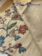 Beige silk cotton saree with colorful Kalamkari-inspired appliqué floral work, elegant woven golden border, handcrafted artistic detailing, soft texture, tassel finish, and graceful traditional drape by Kadamparii Silk.