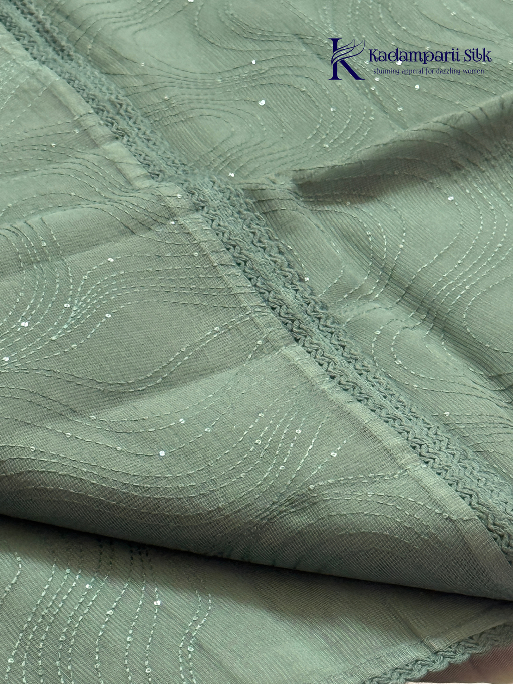 Sage green super net saree with elegant wave-pattern fancy embroidery, subtle shimmer detailing, delicate lace border, lightweight sheer texture, and graceful contemporary drape by Kadamparii Silk.