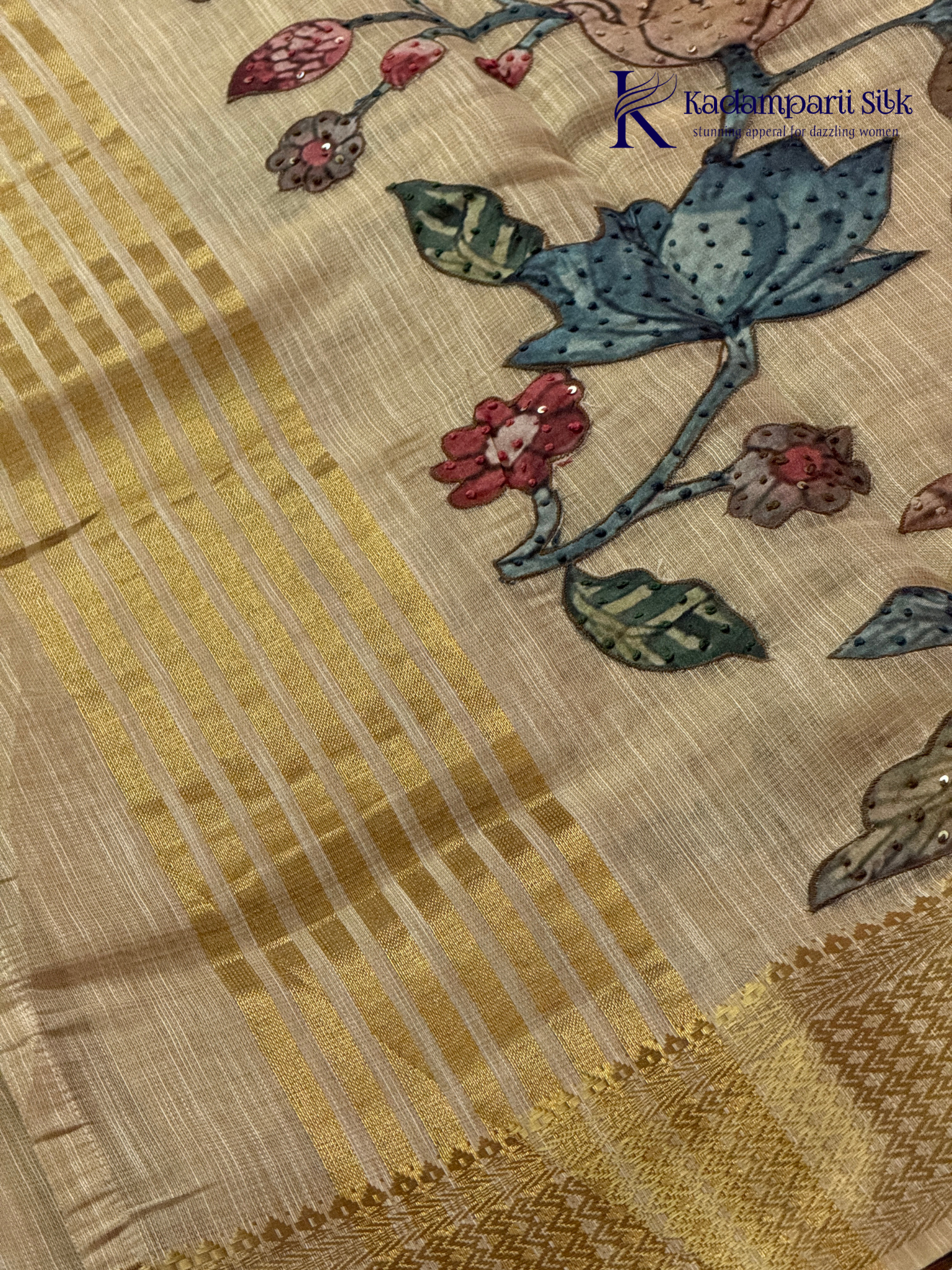 Beige silk cotton saree with colorful Kalamkari-inspired appliqué floral work, elegant woven golden border, handcrafted artistic detailing, soft texture, tassel finish, and graceful traditional drape by Kadamparii Silk.