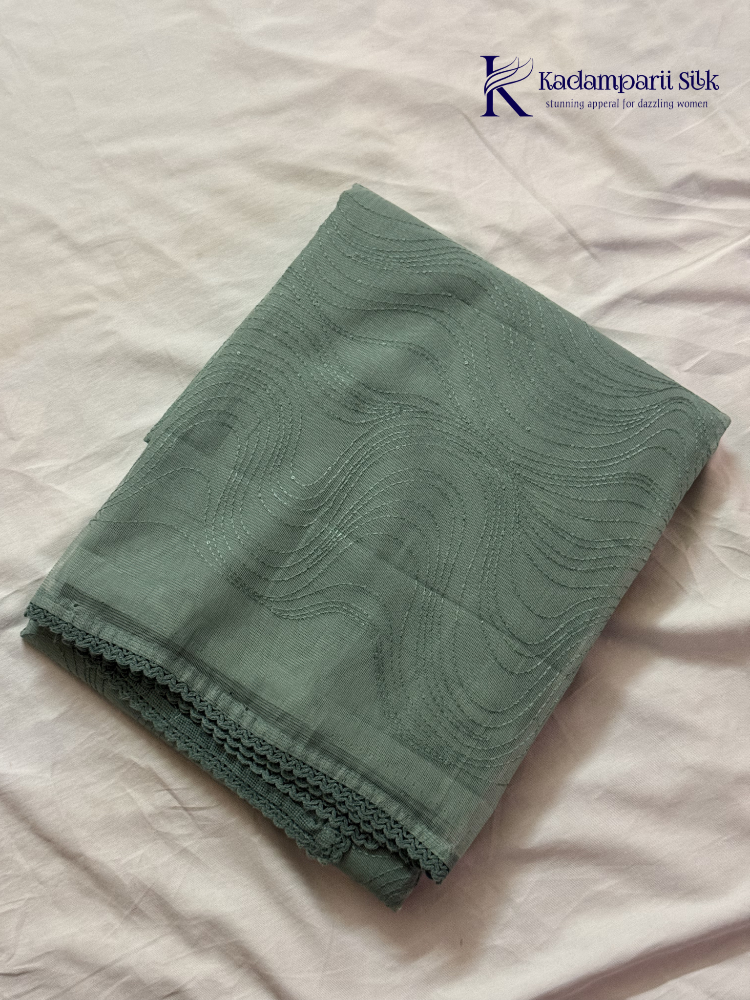 Sage green super net saree with elegant wave-pattern fancy embroidery, subtle shimmer detailing, delicate lace border, lightweight sheer texture, and graceful contemporary drape by Kadamparii Silk.