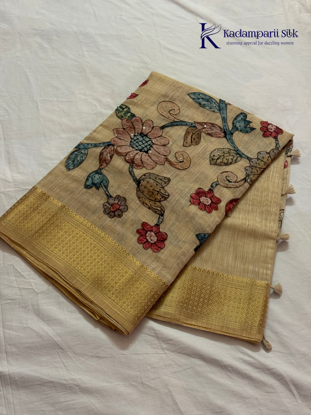 Beige silk cotton saree with colorful Kalamkari-inspired appliqué floral work, elegant woven golden border, handcrafted artistic detailing, soft texture, tassel finish, and graceful traditional drape by Kadamparii Silk.
