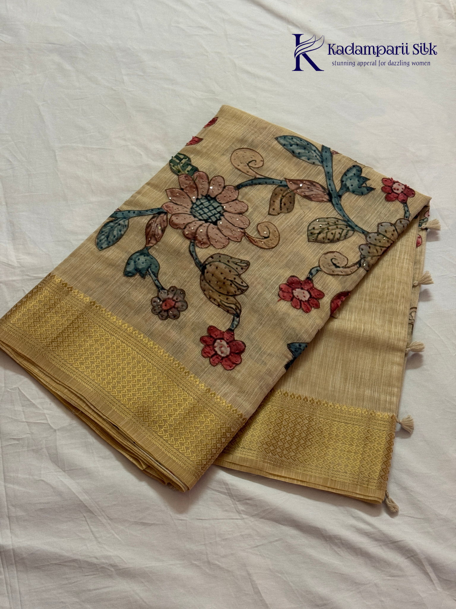 Beige silk cotton saree with colorful Kalamkari-inspired appliqué floral work, elegant woven golden border, handcrafted artistic detailing, soft texture, tassel finish, and graceful traditional drape by Kadamparii Silk.