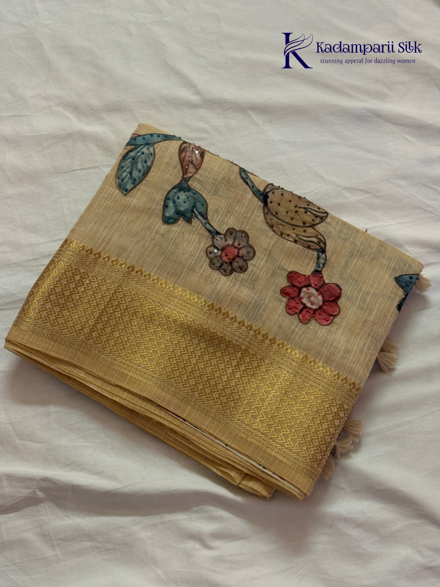 Beige silk cotton saree with colorful Kalamkari-inspired appliqué floral work, elegant woven golden border, handcrafted artistic detailing, soft texture, tassel finish, and graceful traditional drape by Kadamparii Silk.